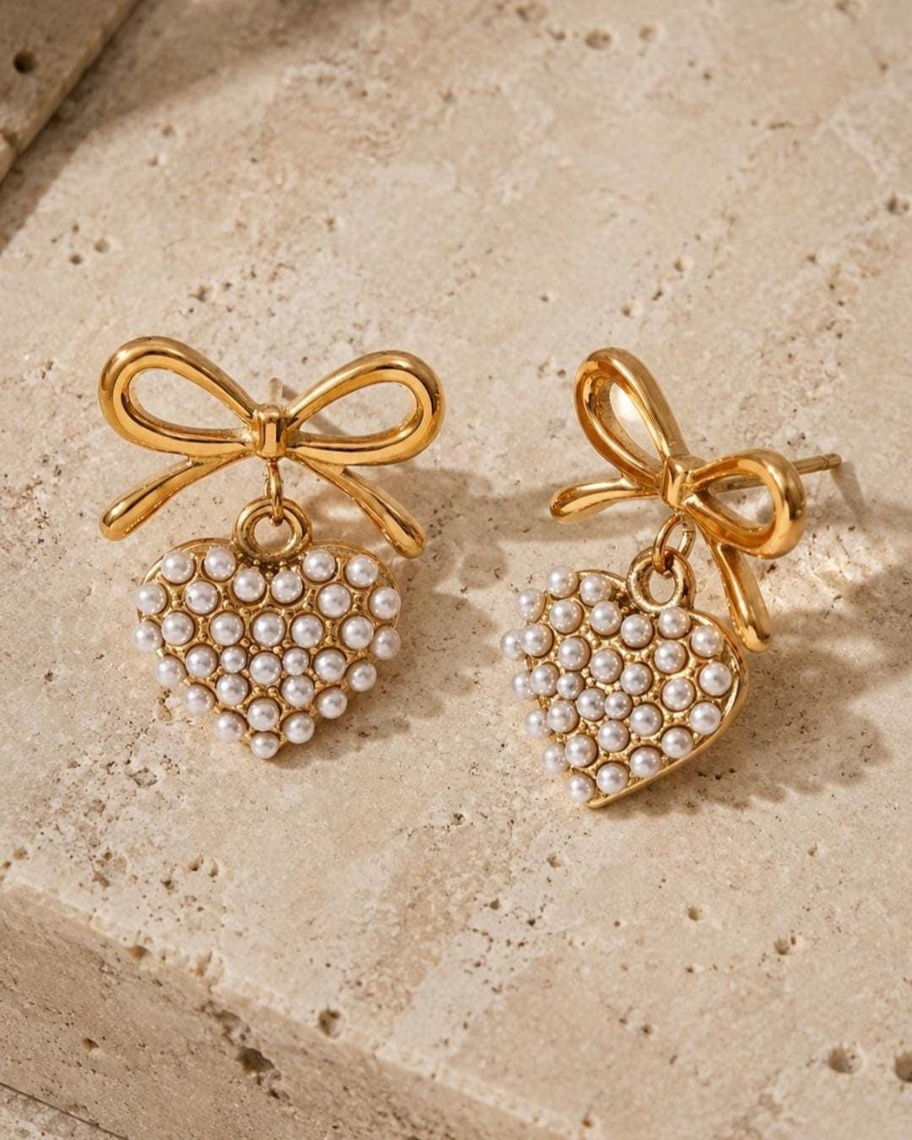 May include: A pair of gold-toned stud earrings featuring a bow design at the top with a dangling heart-shaped charm. The heart charm is filled with small white faux pearls arranged in a dense cluster, creating a textured surface on each earring.
