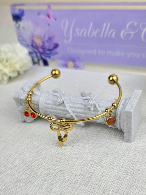 May include: A gold-toned metal cuff bracelet featuring a central bow charm, two small red circular charms on one side, and a single round orange stone charm on the other. The bracelet ends in two smooth gold spheres at the open ends of the band.