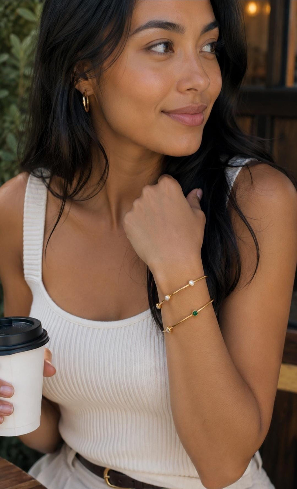 May include: Two thin gold-toned metal bangles worn on the wrist. The top bangle features two small white spherical beads, while the bottom bangle features one small green circular stone and one small white spherical bead set along the band.