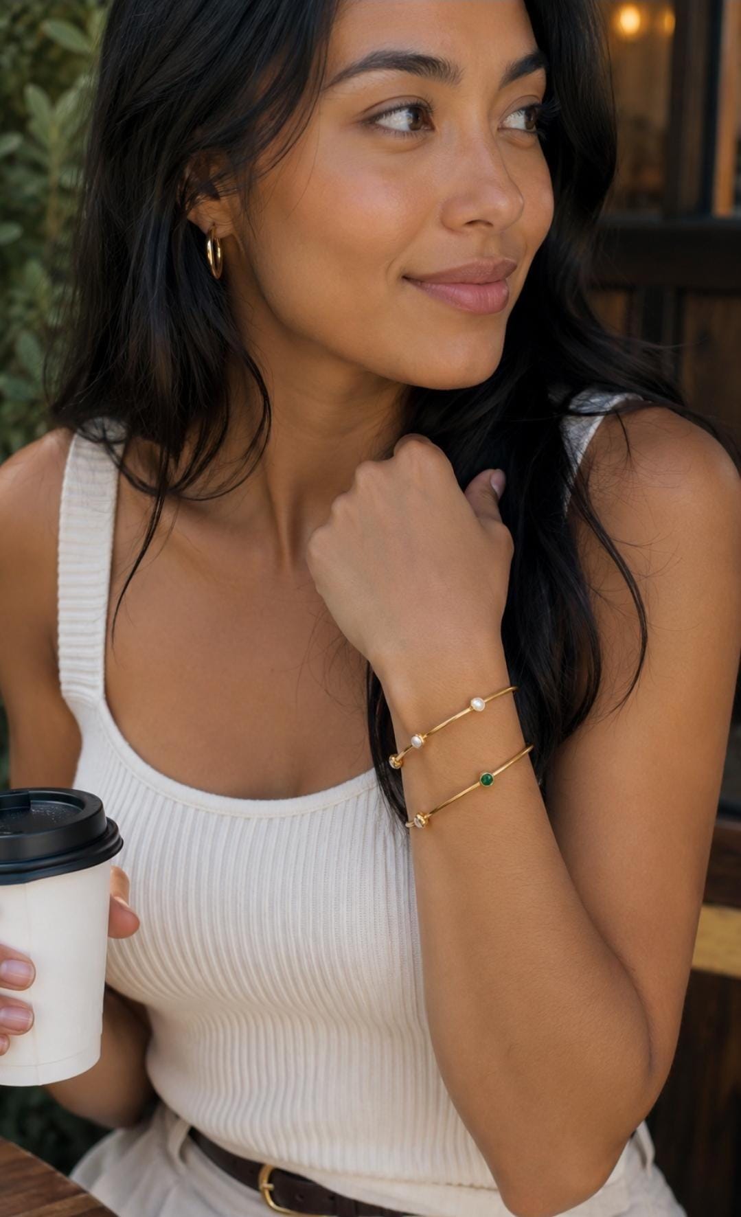 May include: Two thin gold-toned metal bangles worn on the wrist. The top bangle features two small white spherical beads, while the bottom bangle features one small green circular stone and one small white spherical bead set along the band.