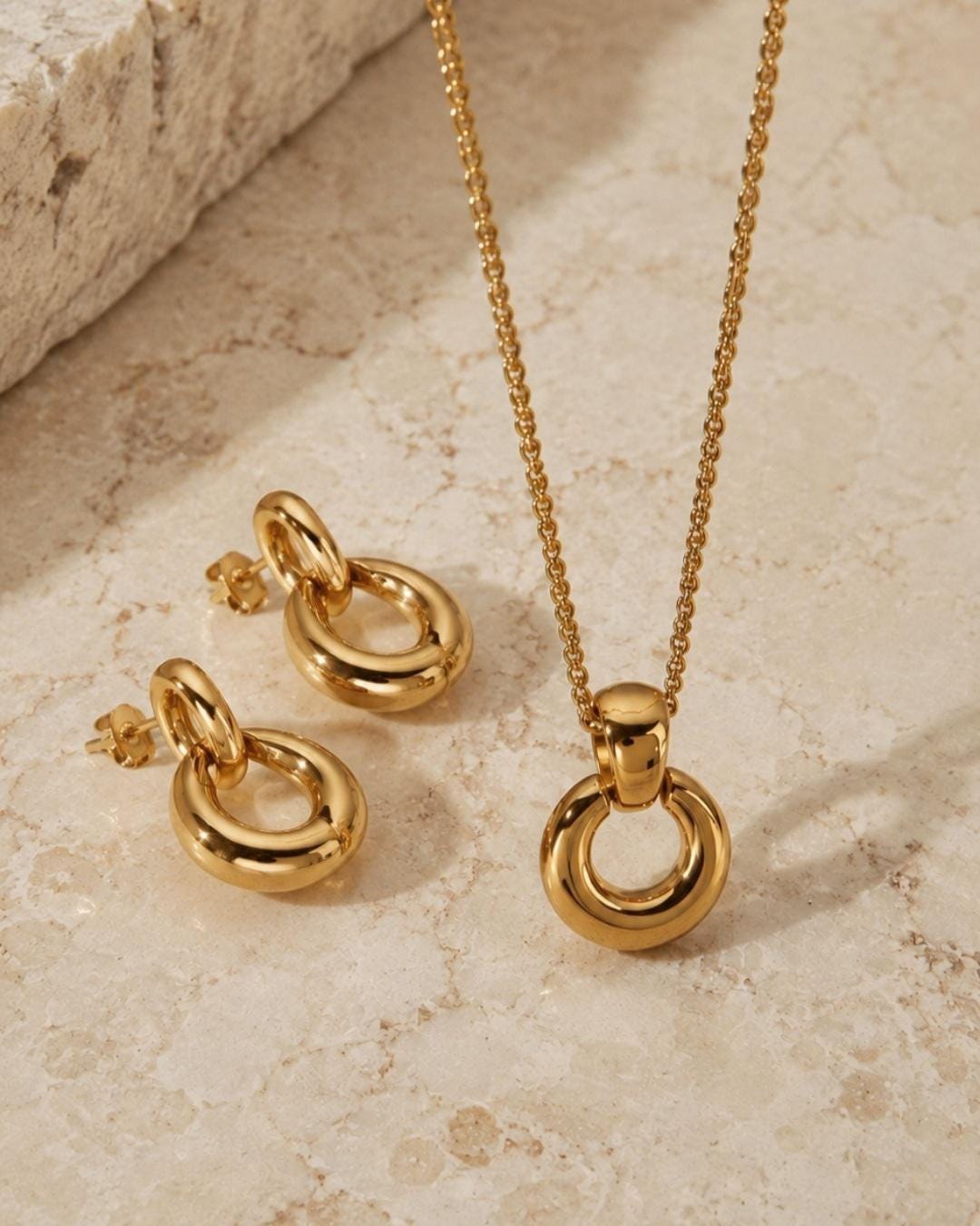 May include: A gold-toned jewelry set consisting of a necklace and a pair of stud earrings. The necklace features a polished circular pendant suspended from a delicate chain. The matching earrings consist of a small hoop attached to a circular charm.